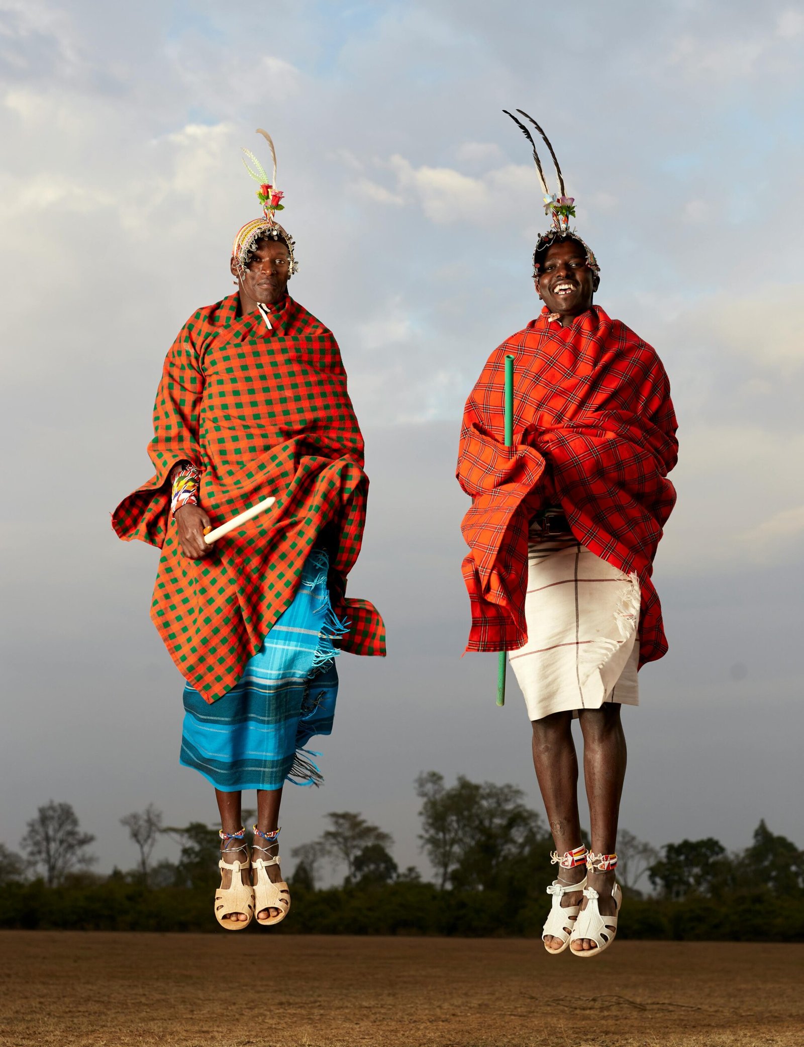 Two Maasai warriors in traditional attire jumping midair in a cloudy field.